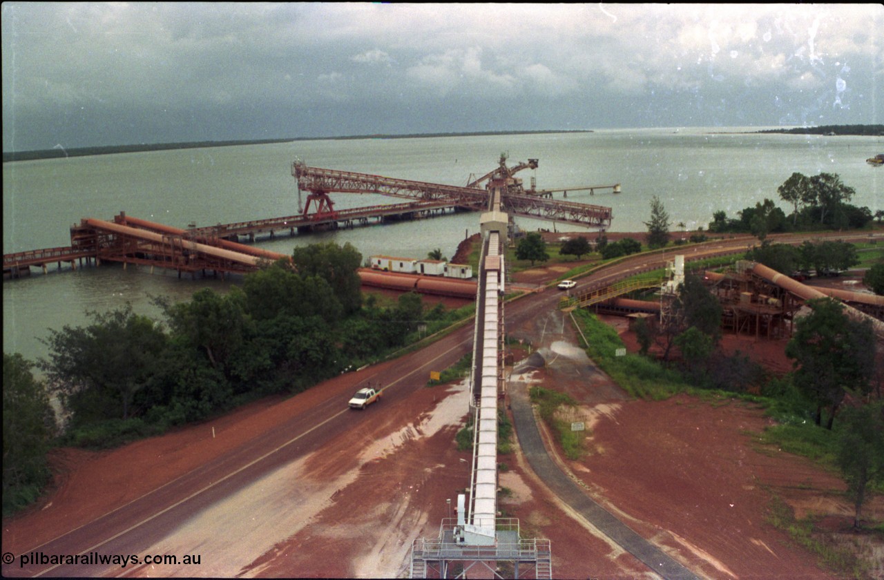211-0A
Weipa, Lorim Point, view from the top of the kaolin storage silos tanks looking north at the kaolin conveyor and ship loader, with red legs and the No.2 bauxite loader straight ahead in the cyclone tie-down position. The bauxite conveyors can be seen passing under the road.
