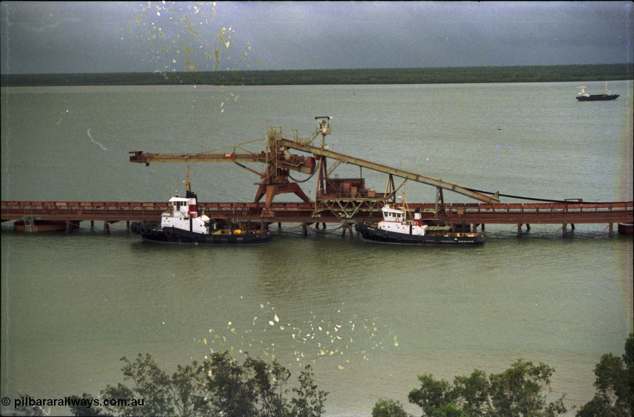 211-01
Weipa, Lorim Point, view from the top of the kaolin storage silos looking west at No.1 bauxite ship loader and the two tug boats. The unloading barge can be seen in the background in the Embley River.
