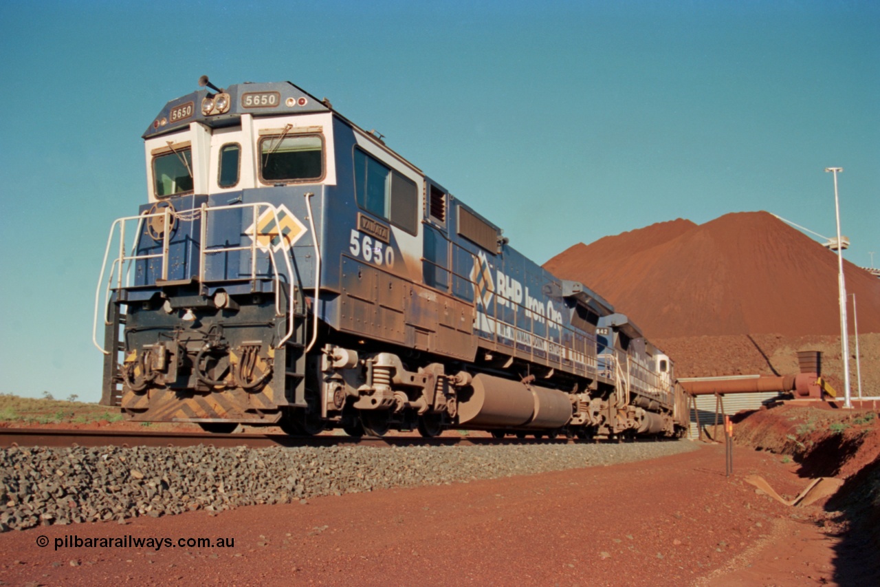 229-22
Yandi Two loadout, looking at the exit portal of the tunnel as Goninan rebuild CM40-8M GE unit 5650 'Yawata' serial 8412-07 / 93-141 drags a loading train through at 1.2 km/h, the massive pile of ore is gravity fed into the waggons via two sets of hydraulic chutes, the original pedestal radial stacker is visible above the ore along with the extraction fan. [url=https://goo.gl/maps/jmtnauf76Zq]GeoData[/url].
Keywords: 5650;Goninan;GE;CM40-8M;8412-07/93-141;rebuild;AE-Goodwin;ALCo;M636C;5481;G6061-2;