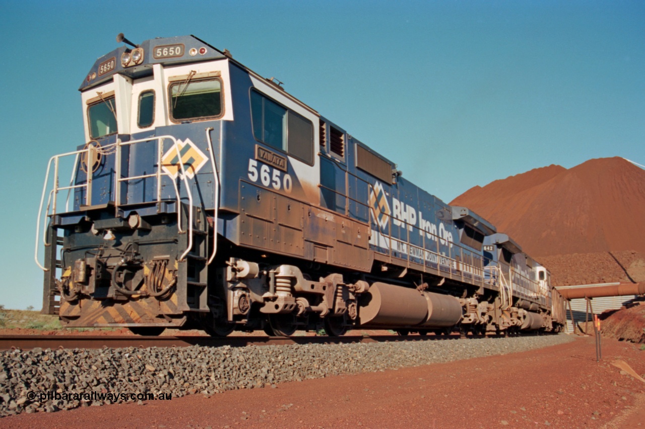 229-23
Yandi Two loadout, looking at the exit portal of the tunnel as Goninan rebuild CM40-8M GE unit 5650 'Yawata' serial 8412-07 / 93-141 drags a loading train through at 1.2 km/h, the massive pile of ore is gravity fed into the waggons via two sets of hydraulic chutes, the original pedestal radial stacker is visible above the ore along with the extraction fan. [url=https://goo.gl/maps/jmtnauf76Zq]GeoData[/url].
Keywords: 5650;Goninan;GE;CM40-8M;8412-07/93-141;rebuild;AE-Goodwin;ALCo;M636C;5481;G6061-2;