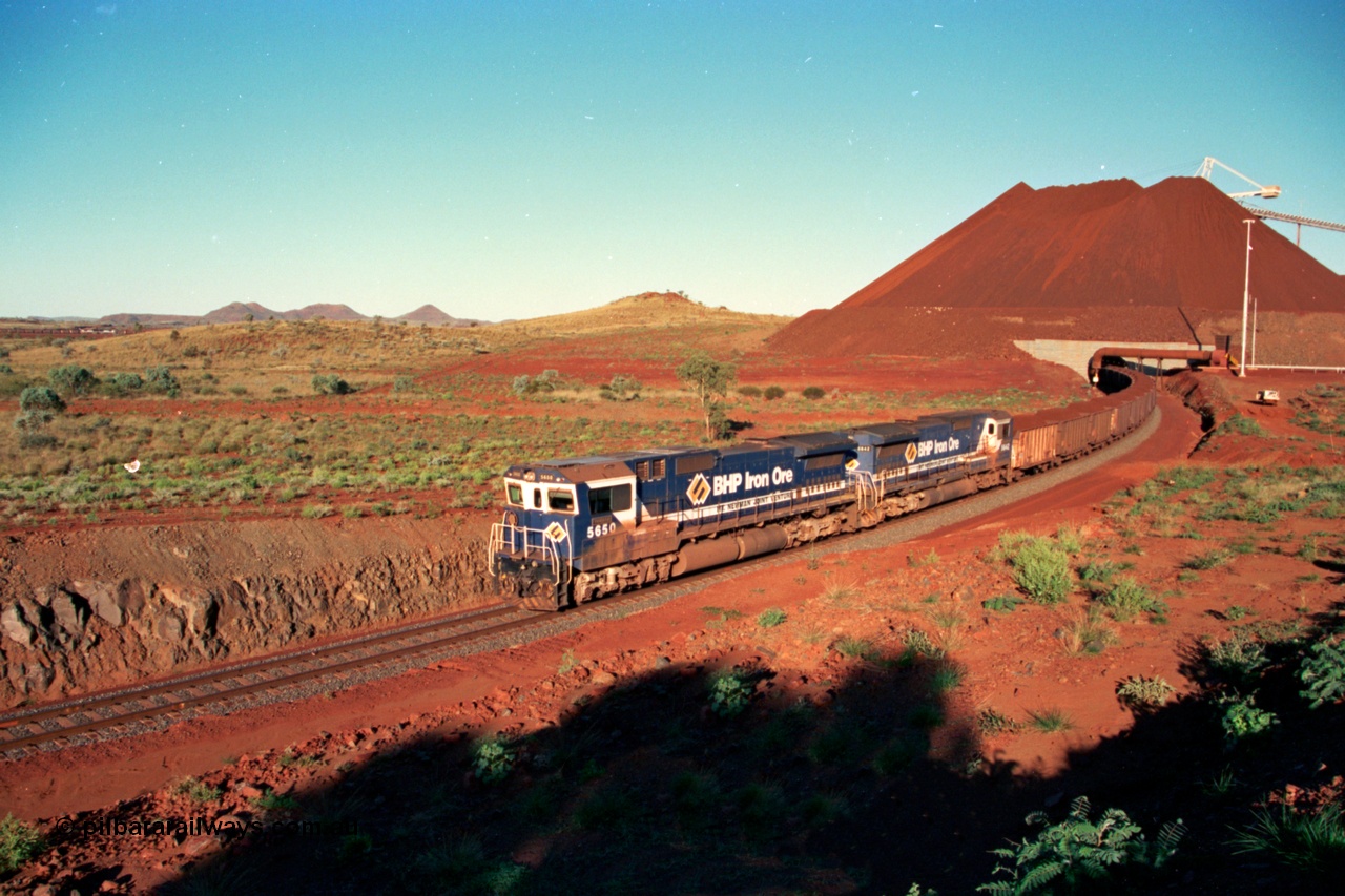 229-30
Yandi Two loadout exit side, as Goninan rebuild CM40-8M GE unit 5650 'Yawata' serial 8412-07 / 93-141 and sister unit drag a loading train at 1.2 km/h, the massive pile of ore is gravity fed into the waggons via two sets of hydraulic chutes, the original pedestal radial stacker is visible above the ore along with the extraction fan, on the left the mid-train remotes can just be made out. [url=https://goo.gl/maps/KQ3dQNrTwd42]GeoData[/url].
Keywords: 5650;Goninan;GE;CM40-8M;8412-07/93-141;rebuild;AE-Goodwin;ALCo;M636C;5481;G6061-2;