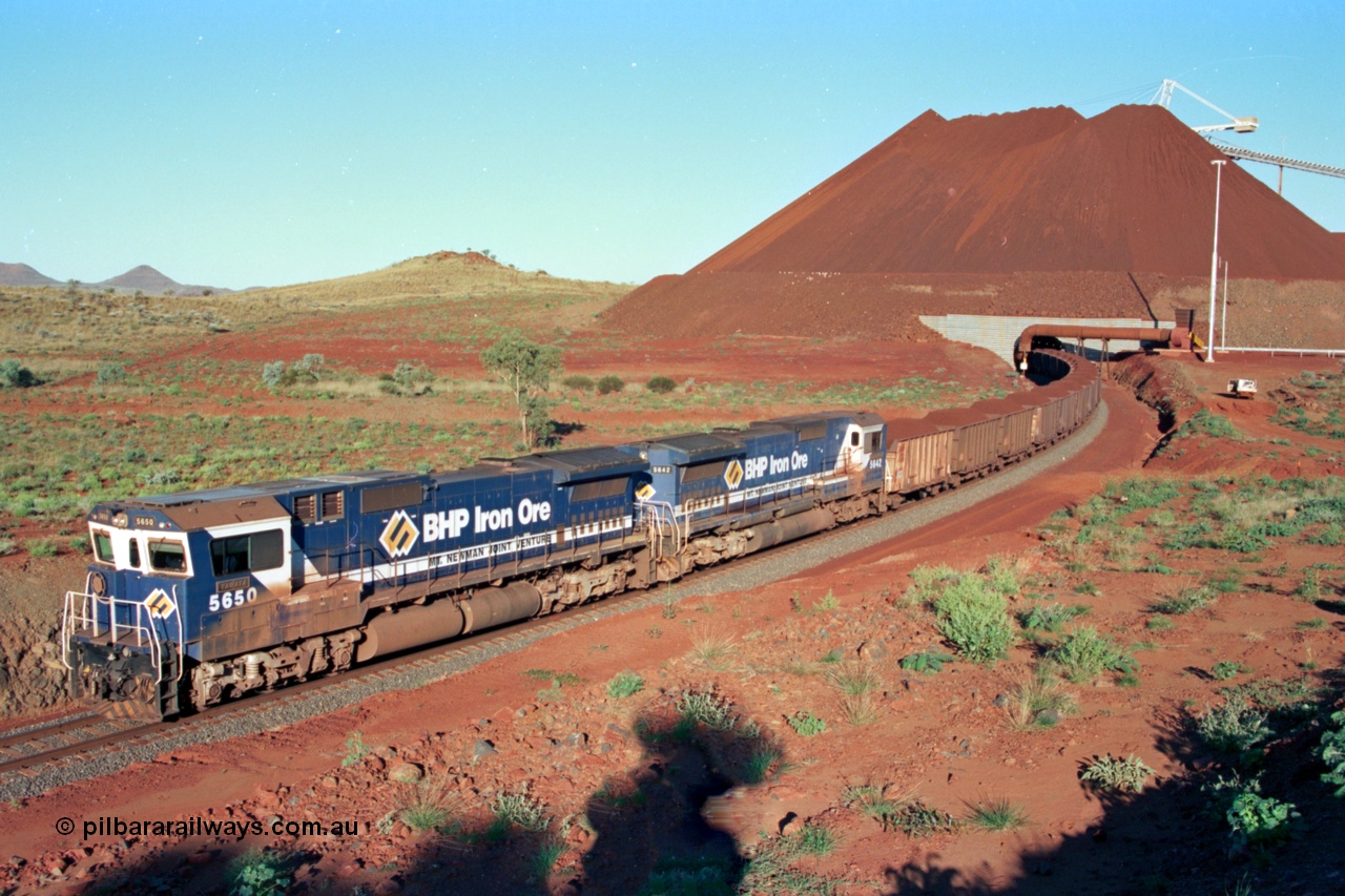 229-31
Yandi Two loadout exit side, as Goninan rebuild CM40-8M GE unit 5650 'Yawata' serial 8412-07 / 93-141 and sister unit 5642 drag a loading train at 1.2 km/h, the massive pile of ore is gravity fed into the waggons via two sets of hydraulic chutes, the original pedestal radial stacker is visible above the ore along with the extraction fan, to the left in the distance is a partial glimpse of the Three Sisters. [url=https://goo.gl/maps/KQ3dQNrTwd42]GeoData[/url].
Keywords: 5650;Goninan;GE;CM40-8M;8412-07/93-141;rebuild;AE-Goodwin;ALCo;M636C;5481;G6061-2;