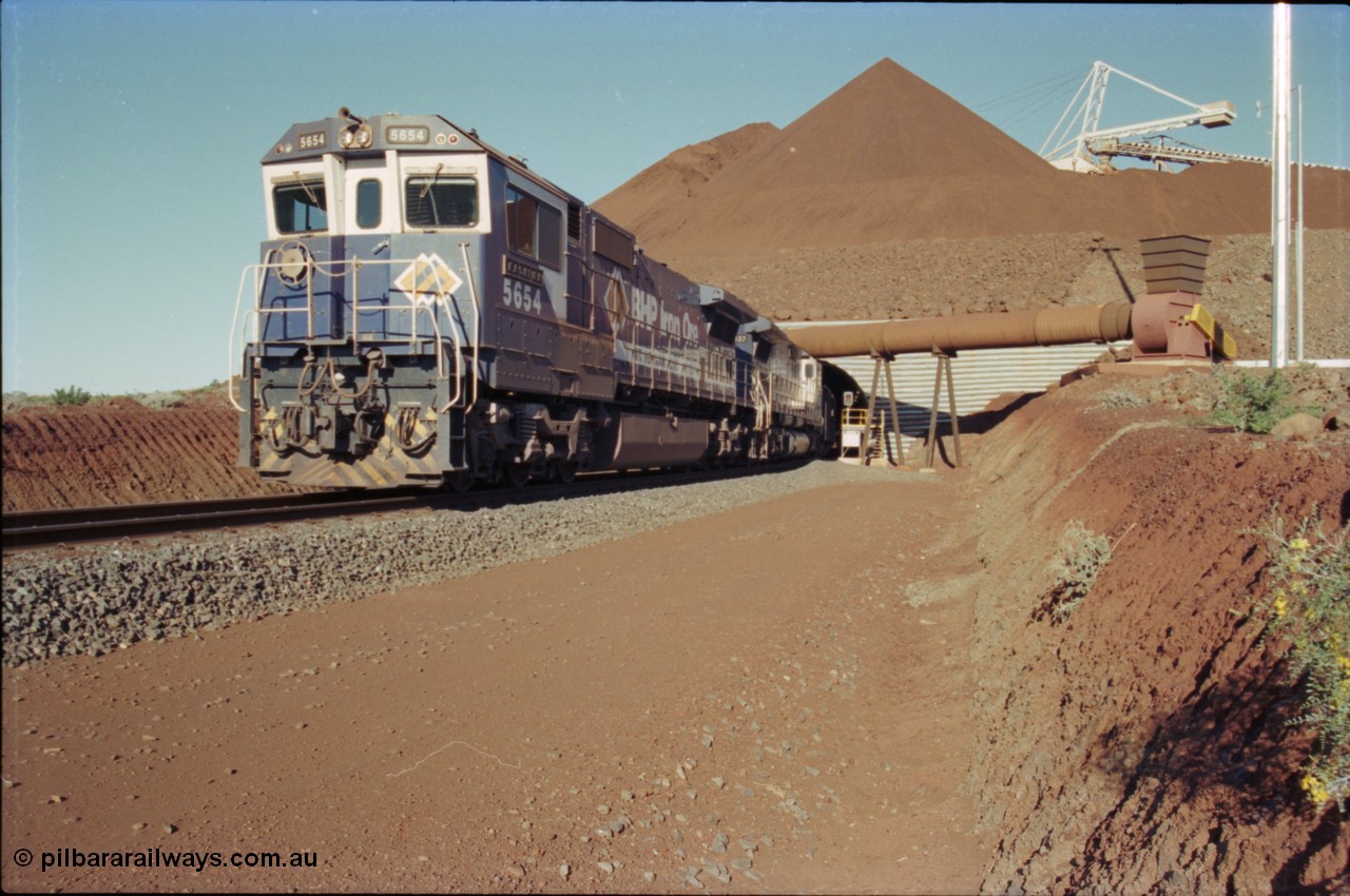 230-19
Yandi Two load-out tunnel exit side portal, Goninan rebuild GE CM40-8M model 5654 'Kashima' serial 8412-11/93-145 leads a loading train 1.2 km/h. The ore stockpile and pedestal stacker are visible. The pipe and fan are for the dust extraction system. The livery is the marigold version of the BHP Australia logo. Geodata [url=https://goo.gl/maps/h68ia1caJvL2] -22.718338, 119.039909 [/url].
Keywords: 5654;Goninan;GE;CM40-8M;8412-11/93-145;rebuild;Comeng-NSW;ALCo;M636C;5493;C6084-9;