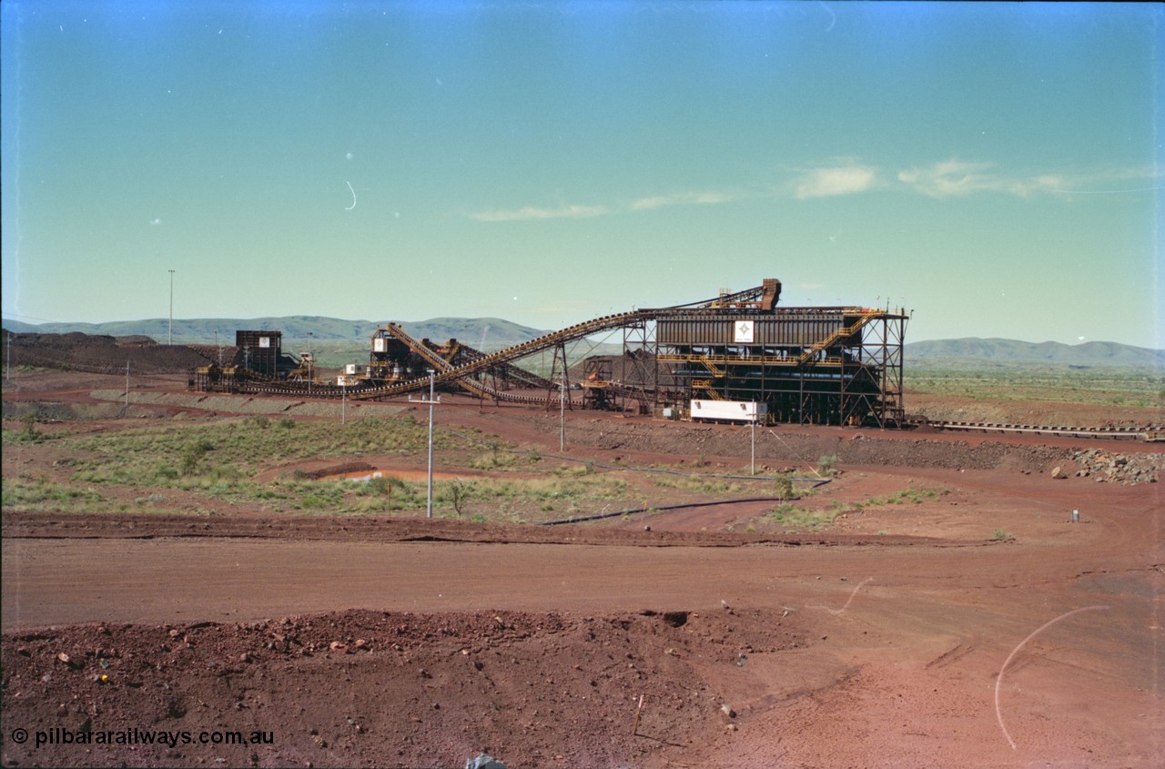 232-00
Yandi Two crushing and screening plant, built and operated by Henry Walker for BHP Iron Ore, overview from the radial stacker feed conveyor. From the left is the primary crusher, the dual secondary crushers can be made out behind the tertiary crushers in the middle and the screenhouse is the large building tot he right, the conveyor leading to the right is CV210. February 1997. [url=https://goo.gl/maps/TUReAkezgfz]GeoData[/url].
