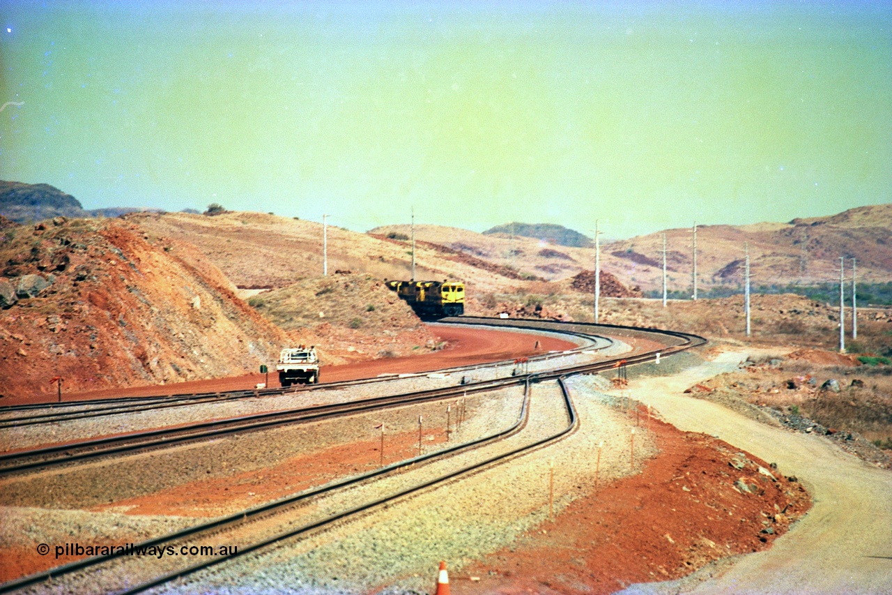 246-06
Cape Lambert, yard view of the then new extension to the south for the West Angelas mine was coming on stream as a Robe River loaded Deepdale train arrives on the main behind the standard quad Dash 8 power with 202 waggons, the just visible siding (rejoining) on the far left will become a compressor waggon holding road, the 6 km and Boat Beach Rd grade crossing are around the corner to the left. Closest to the camera is the extended No. 1 Road which becomes the Empty Car Line from car dumper one for the Deepdale traffic. Next is the original line or Mainline and the third line across is the Loaded Car Line for the West Angelas traffic to the new car dumper 2. 22nd May 2002.
