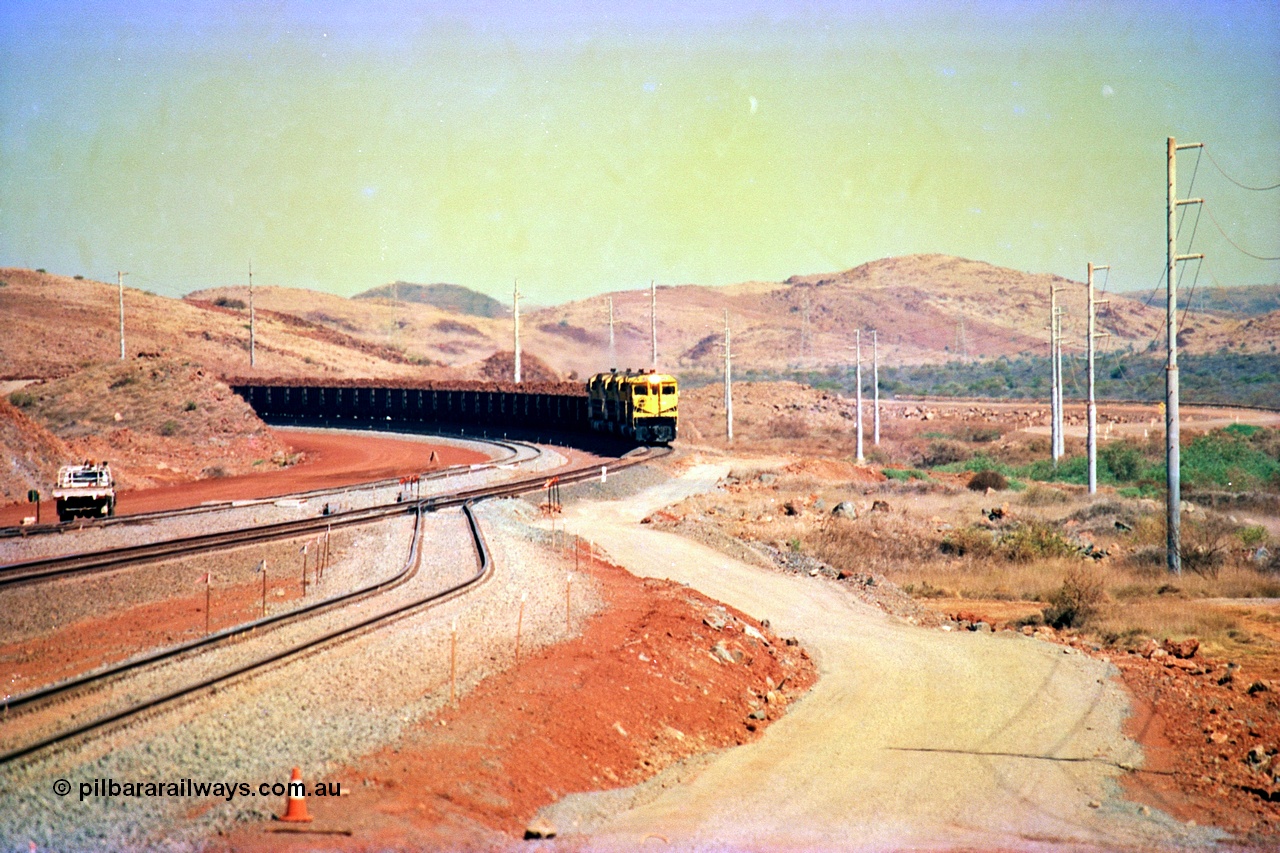 246-08
Cape Lambert, yard view of the then new extension to the south for the West Angelas mine was coming on stream as a Robe River loaded Deepdale train arrives on the main behind the standard quad Dash 8 power with 202 waggons, the just visible siding (rejoining) on the far left will become a compressor waggon holding road, the 6 km and Boat Beach Rd grade crossing are around the corner to the left. Closest to the camera is the extended No. 1 Road which becomes the Empty Car Line from car dumper one for the Deepdale traffic. Next is the original line or Mainline and the third line across is the Loaded Car Line for the West Angelas traffic to the new car dumper 2. 22nd May 2002.
