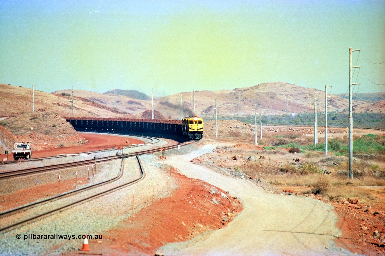 246-09
Cape Lambert, yard view of the then new extension to the south for the West Angelas mine was coming on stream as a Robe River loaded Deepdale train arrives on the main behind the standard quad Dash 8 power with 202 waggons, the just visible siding (rejoining) on the far left will become a compressor waggon holding road, the 6 km and Boat Beach Rd grade crossing are around the corner to the left. Closest to the camera is the extended No. 1 Road which becomes the Empty Car Line from car dumper one for the Deepdale traffic. Next is the original line or Mainline and the third line across is the Loaded Car Line for the West Angelas traffic to the new car dumper 2. 22nd May 2002.
