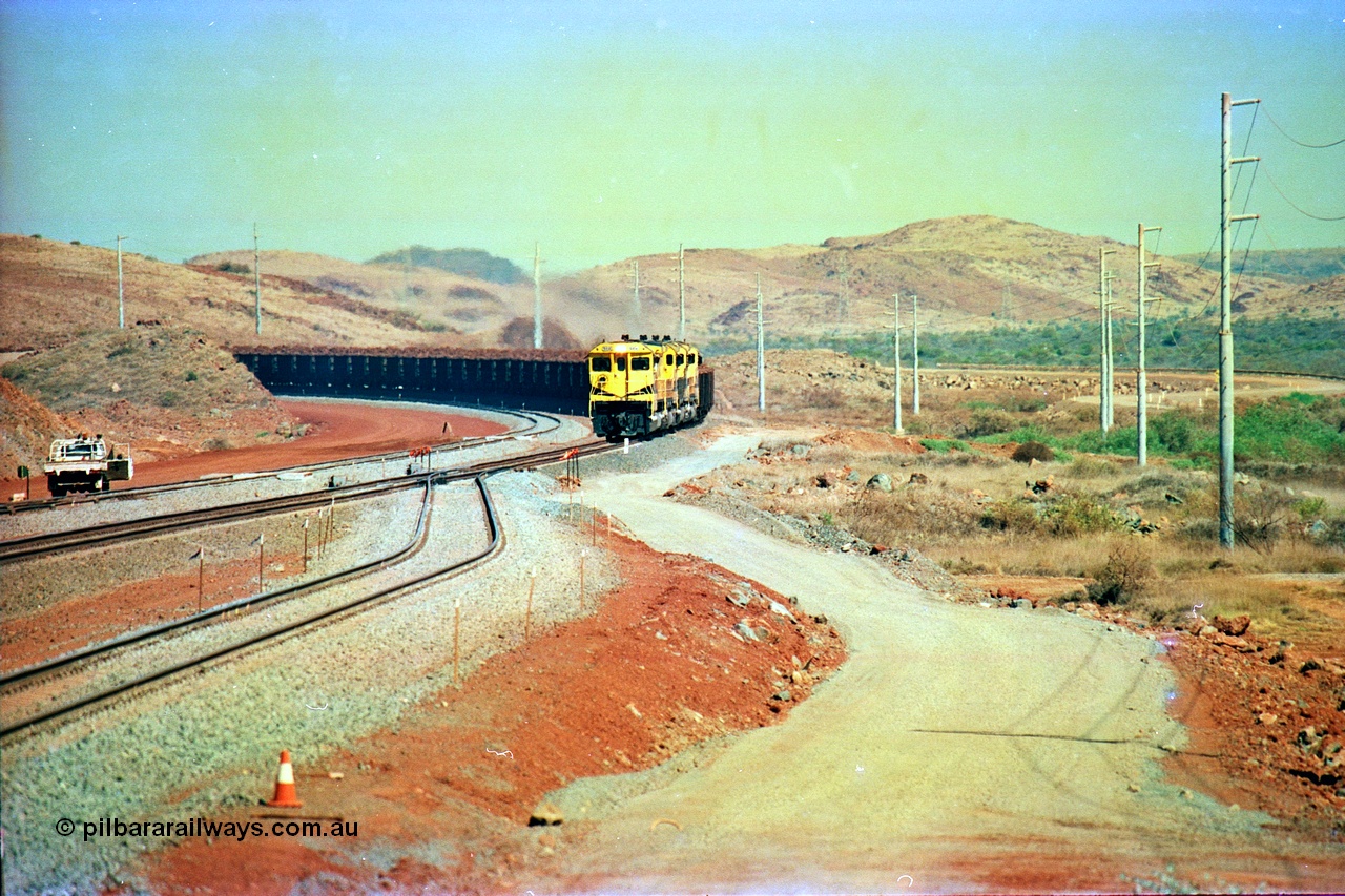 246-10
Cape Lambert, yard view of the then new extension to the south for the West Angelas mine was coming on stream as a Robe River loaded Deepdale train arrives on the main behind the standard quad Dash 8 power with 202 waggons, the just visible siding (rejoining) on the far left will become a compressor waggon holding road, the 6 km and Boat Beach Rd grade crossing are around the corner to the left. Closest to the camera is the extended No. 1 Road which becomes the Empty Car Line from car dumper one for the Deepdale traffic. Next is the original line or Mainline and the third line across is the Loaded Car Line for the West Angelas traffic to the new car dumper 2. 22nd May 2002.
