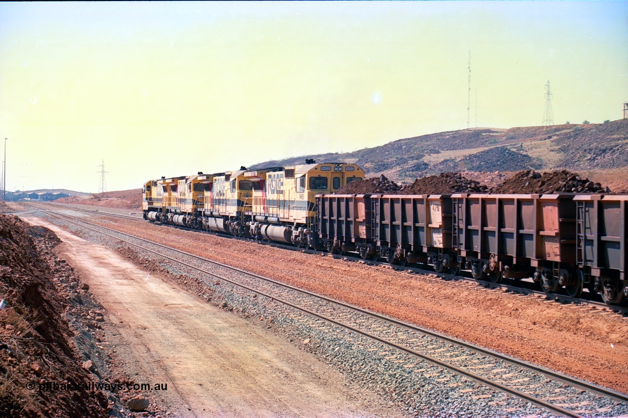 246-27
Cape Lambert, a Robe River loaded Deepdale train rolls along the mainline behind the standard quad Dash 8 power with 202 waggons with newly extended roads either side of it. In the distance can be seen are the railway workshops. 22nd May 2002.
