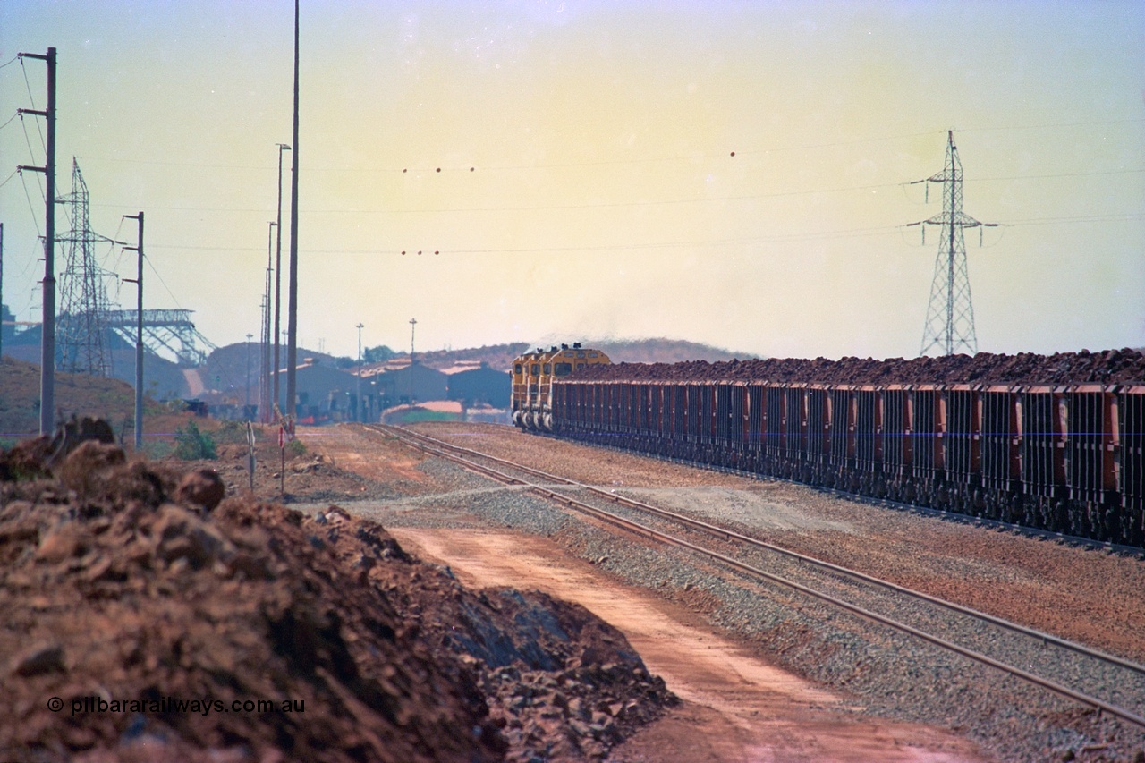 246-36
Cape Lambert, a Robe River loaded Deepdale train rolls along the mainline behind the standard quad Dash 8 power with 202 waggons which are a mix of builds from Nippon Sharyo Japan and Tomlinson Steel and Centurion Industries both WA companies. The ore is a ROM product and will be dumped over the gyratory crusher to be correctly sized. The newly extended roads can be seen either side of the train and in the distance can be seen are the railway workshops. 22nd May 2002.
