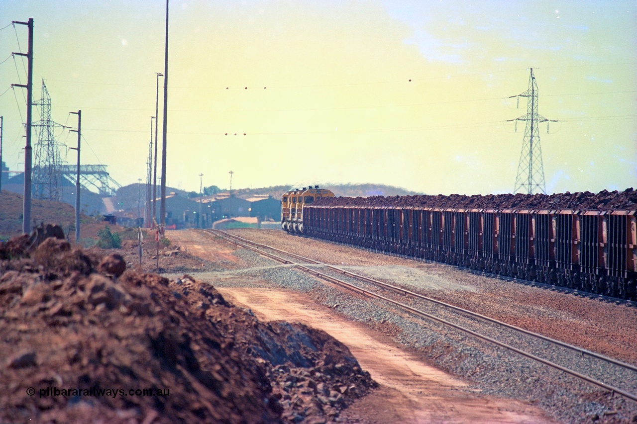 246-37
Cape Lambert, a Robe River loaded Deepdale train rolls along the mainline behind the standard quad Dash 8 power with 202 waggons which are a mix of builds from Nippon Sharyo Japan and Tomlinson Steel and Centurion Industries both WA companies. The ore is a ROM product and will be dumped over the gyratory crusher to be correctly sized. The newly extended roads can be seen either side of the train and in the distance can be seen are the railway workshops. 22nd May 2002.
