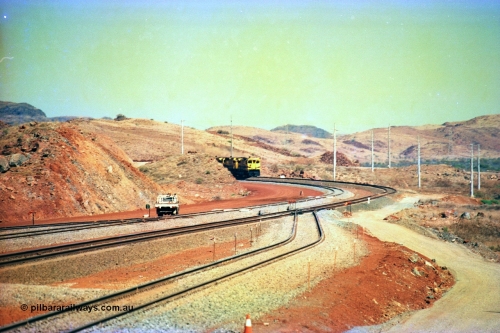 246-06
Cape Lambert, yard view of the then new extension to the south for the West Angelas mine was coming on stream as a Robe River loaded Deepdale train arrives on the main behind the standard quad Dash 8 power with 202 waggons, the just visible siding (rejoining) on the far left will become a compressor waggon holding road, the 6 km and Boat Beach Rd grade crossing are around the corner to the left. Closest to the camera is the extended No. 1 Road which becomes the Empty Car Line from car dumper one for the Deepdale traffic. Next is the original line or Mainline and the third line across is the Loaded Car Line for the West Angelas traffic to the new car dumper 2. 22nd May 2002.
