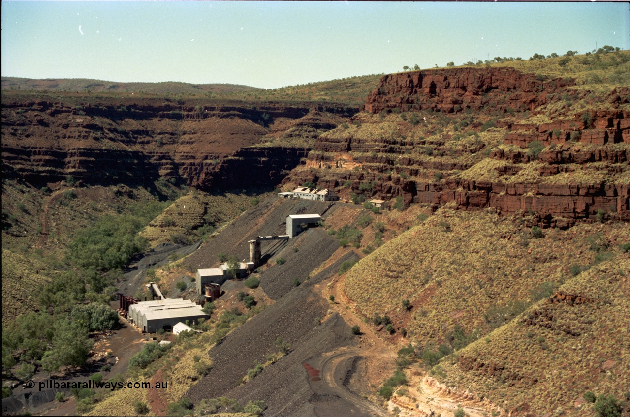 195-16
Wittenoom, view from the Devil's Staircase of Colonial Mill and Mine with piles of tailings, milling site overview, looking south, railway workshops and underground offices are the higher buildings, then the primary crushing shed, holding silo, down to the mill and then the bag house and associated sheds, tailings line the gorge side.
