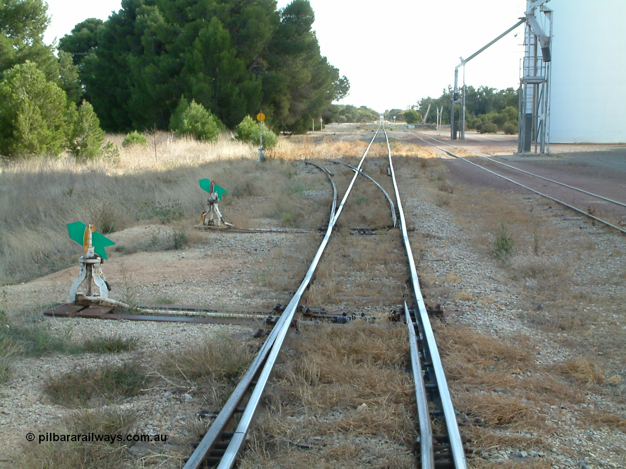 030406 092313
Yeelanna, station yard view looking north down the mainline from the south end of the station siding, points, leaver and indicator for the Kapinnie and former Mount Hope line at the bottom of frame, then the south end points, lever and indicator for the station siding, and then the high yellow disk for the southern leg of the triangle can be seen. With silo outflow spouts on the right. 6th April 2003.
