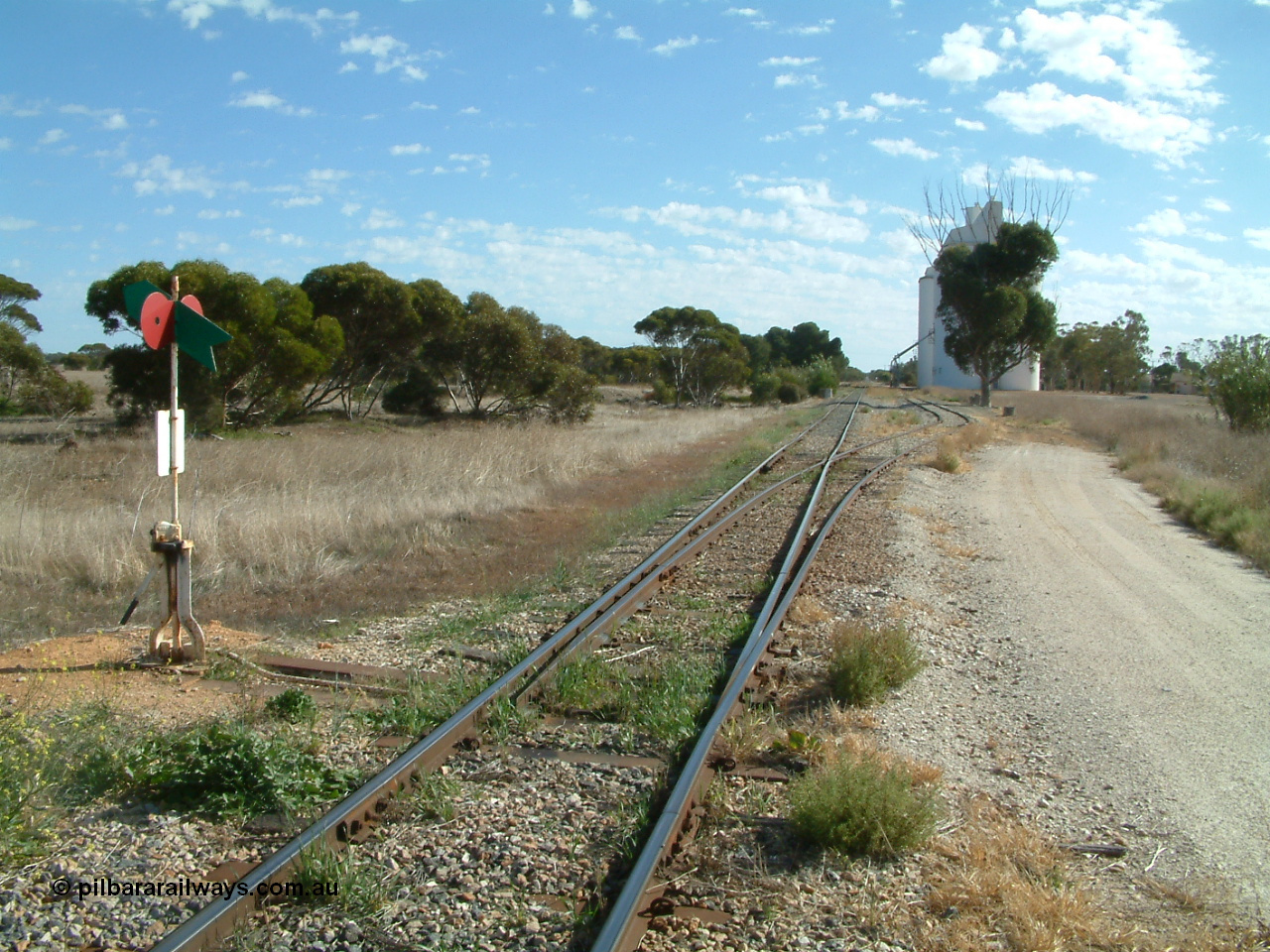 030406 093000
Yeelanna, station located at the 81.7 km, original terminus of line in April 1909, then junction for line to Mount Hope in October 1914. Station yard overview from the south end looking north, points, lever and indicator for the grain siding with concrete silo complexes in the background. The branch line to Kapinnie and former Mount Hope is just visible through the trees on the raised embankment. 6th April 2003.
