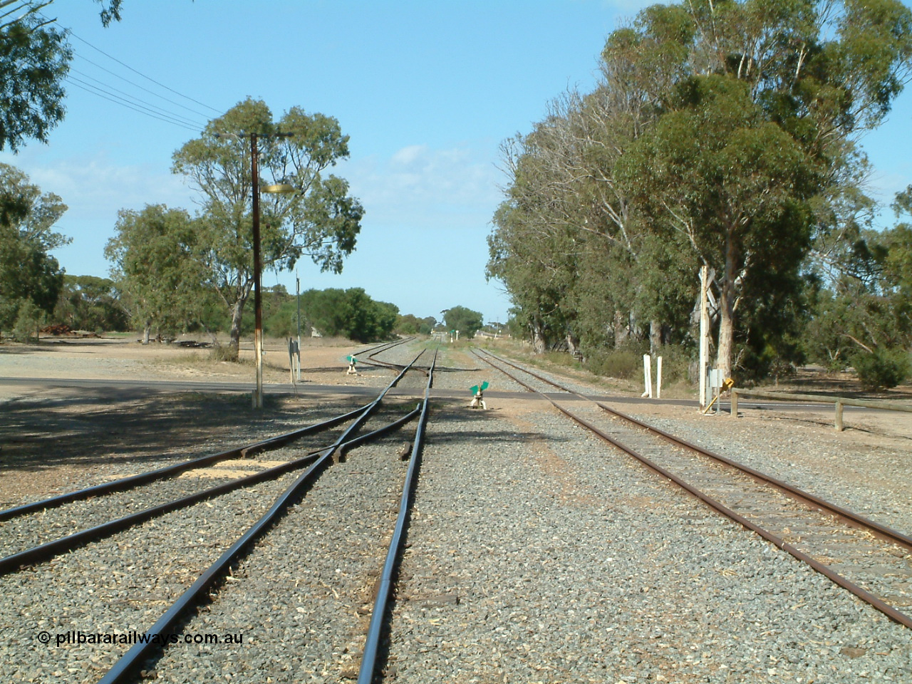 030406 100006
Cummins, yard view looking south across Railway Tce grade crossing, Buckleboo line - siding converging on the left, with the grain siding on the right, the points, indicator and lever south of the grade crossing is to the grain bunker loading - unloading site which is behind the trees to the left. 6th April 2003.
