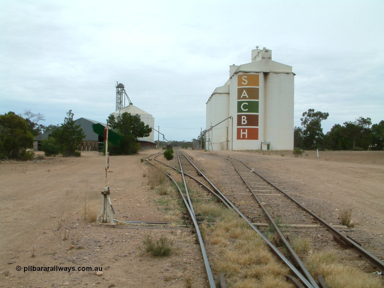 030407 104705
Poochera, station yard overview looking north from south end of Goods Siding 2, point lever and indicator for siding, on the left is the horizontal grain shed with an Ascom style silo complex behind it, on the right the goods shed can be made out along with the SACBH concrete silo complexes. 7th April 2003.
