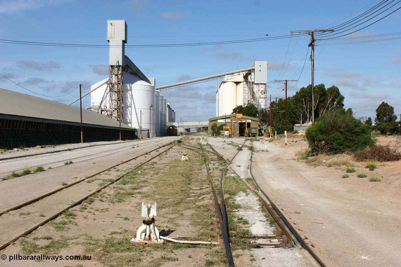 060113 2459
Thevenard, yard view looking west from No. 13 points at the front of the maintenance 'yard' with the disused grain bunker and 6DD3 running along beside the silos as it unloads at GRA, further silos form the background with the maintenance centre with stand-by loco 871 on hand and a couple of ENH hopper waggons. 13th January 2006.
