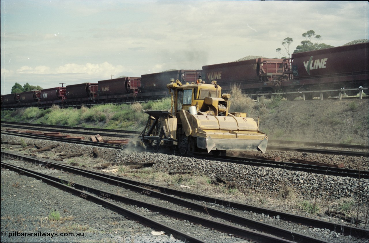 125-10
Seymour, standard gauge mainline, ballast regulator working past loco depot, new and old sleepers along line, broad gauge ballast rake in background.
