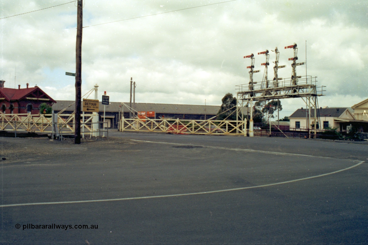 139-14
Ballarat, view of Lydiard Street interlocked crossing gates and semaphore signal bridge, signal box is just to the left of frame, and station is to the right. Ballarat Coachlines on the brick building on the left, entrance to Ballarat Freightgate in middle of frame at V/Line sign.
