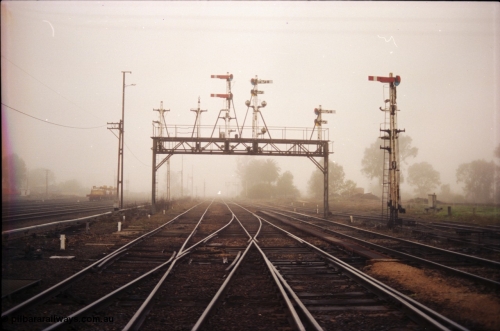 142-1-14
Benalla station yard overview looking north, signal gantry with disc posts 23 and 24 stripped and 25 partially stripped, interlocking crossing under tracks, signal post 27 partially stripped, Yarrawonga line double compound points in middle of frame, very foggy.
