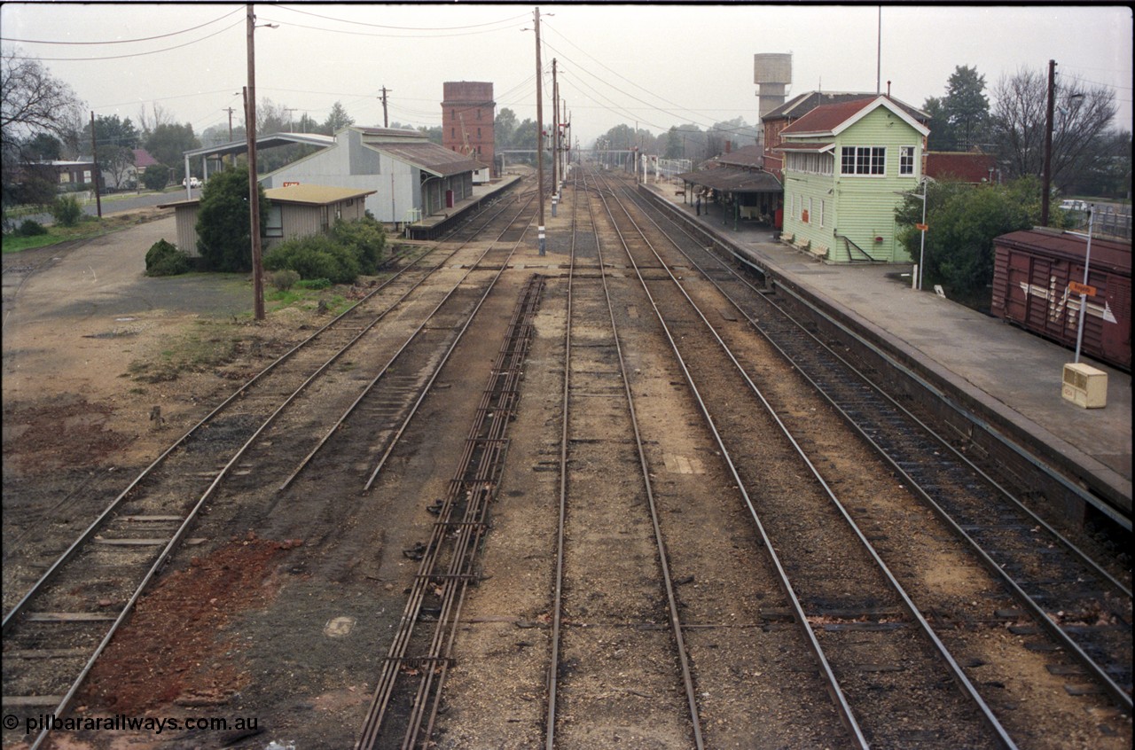 142-2-01
Wangaratta station yard overview looking north from footbridge, No.4 Rd disconnected, goods shed and platform and Freightgate canopy, water tower and crane on the left, station building and elevated signal box on the right, north end footbridge visible in the distance, point rodding running down middle of yard.
