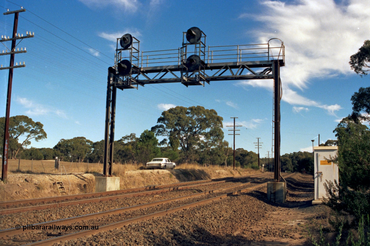 153-2-04
Bank Box Loop, broad gauge track view of signal gantry at Ballarat end, searchlight down home signals 32 for loop departures and 30 for mainline departures, looking towards Ballarat, train control phone booth at right, HK Holden framed under gantry.
