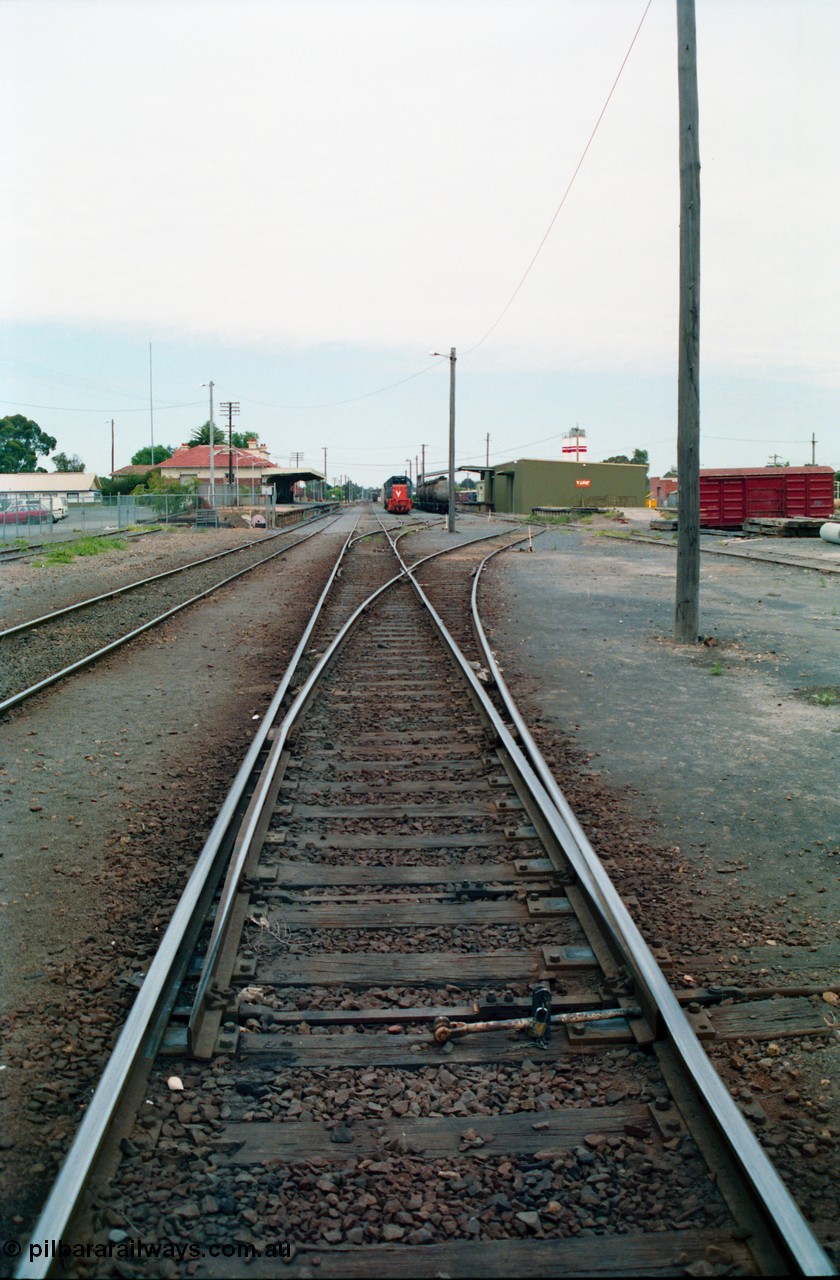 156-08
Shepparton yard view looking south, points for No.5 Road off No.2 Road, then points for No.3 and 4 Roads, engine siding at left, and No.1 Road running down to station platform and building, grounded B van at right, with stabled T class locomotive and fuel waggons with goods shed and Freightgate and cement silos in the background.
