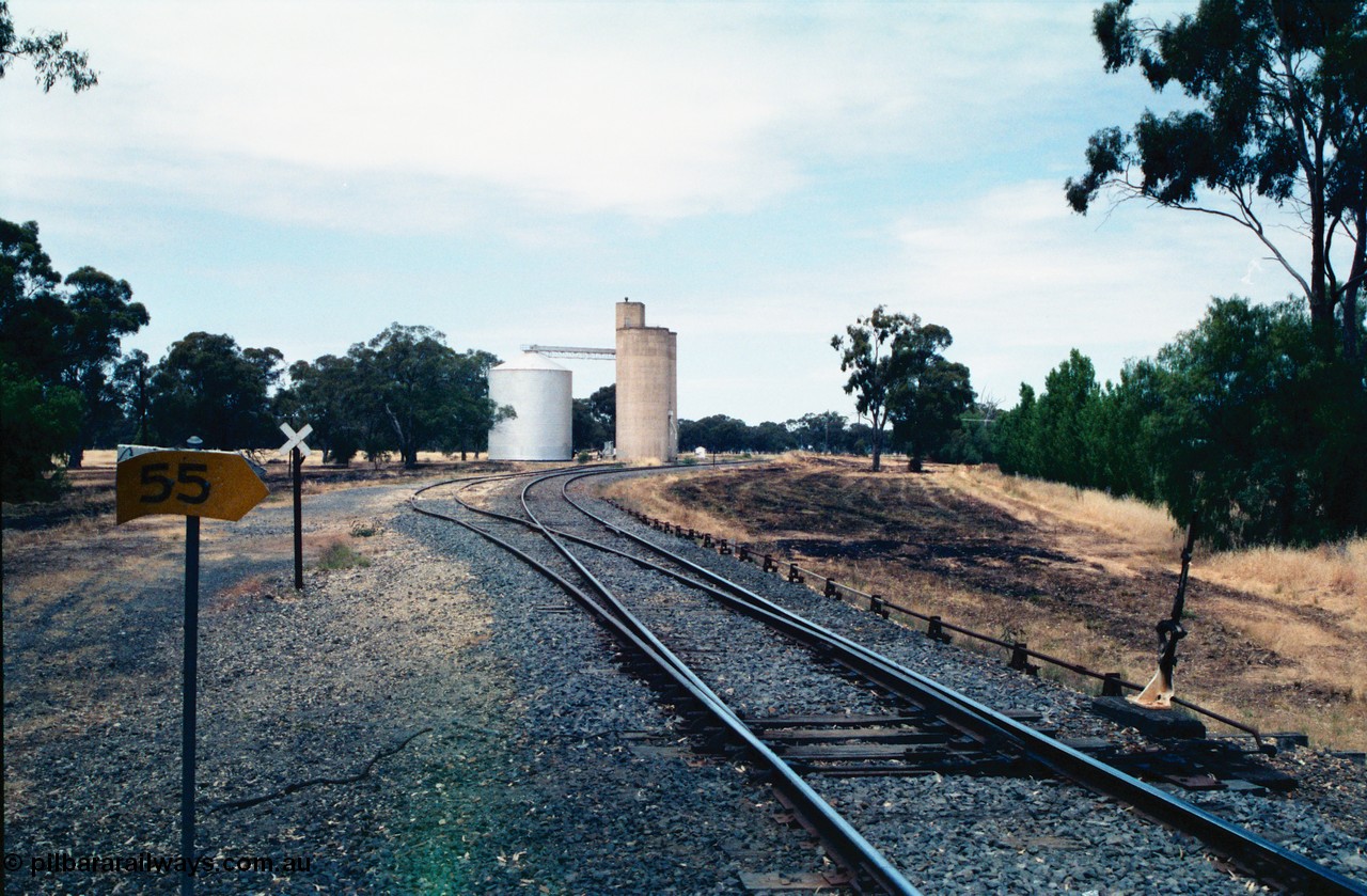 156-20
Wunghnu, station yard overview, looking south, mainline points to siding, lever, interlocking and point rodding for siding derail, Williamstown style silo complex with steel annex, yellow 55 km/h speed board and white cross grade crossing indicator signs.
