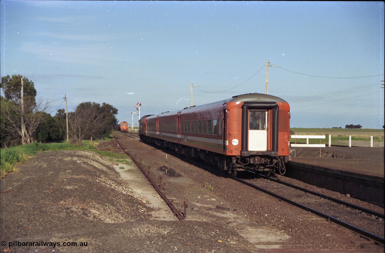 157-02
Gheringhap, station view looking east from former up platform, V/Line broad gauge A class A 77 Clyde Engineering EMD model AAT22C-2R serial 83-1181 rebuilt from B 77 Clyde Engineering EMD model ML2 serial ML2-18 leads the up Ararat to Melbourne via Nth Geelong passenger train with a three car N set, along the mainline heading now for North Geelong, a down grain train 9121 sits in Siding A, station platform, point rodding and semaphore signals.
