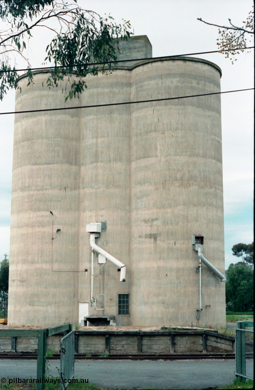 161-03
Numurkah, view from outside platform looking across yard through gates, goods loading platform visible with Williamstown style silo complex located behind it with train loading spouts.
