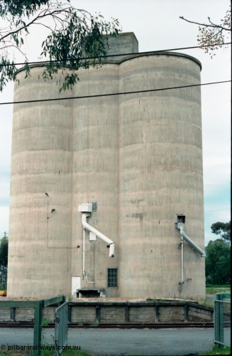161-03
Numurkah, view from outside platform looking across yard through gates, goods loading platform visible with Williamstown style silo complex located behind it with train loading spouts.
